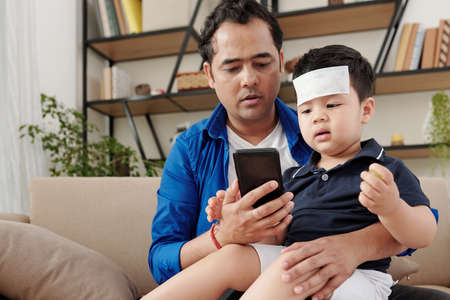 Worried father sitting on sofa with sick child on his laps and calling ambulanceの写真素材