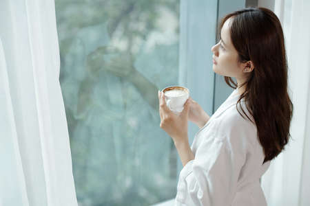Pensive young woman drinking cup of morning coffee and looking outside through apartment windowの写真素材