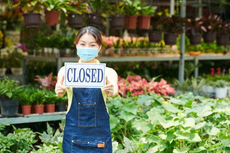Young woman in protective mask standing in gardening center and showing closed sign