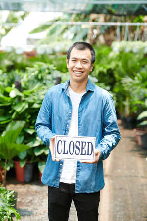 Cheerful young man holding closed sign when standing in gardening centerの写真素材