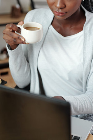 Cropped image of young woman with full lips drinking cup of coffee and working on laptopの写真素材