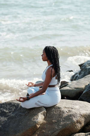 Calm woman in actvewear enjoying sound of waves when sitting in lotus postion and meditating with eyes closedの写真素材