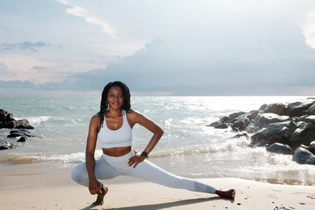 Smiling pretty sportswoman doing deep lunges when exercising on sandy beach after jogging