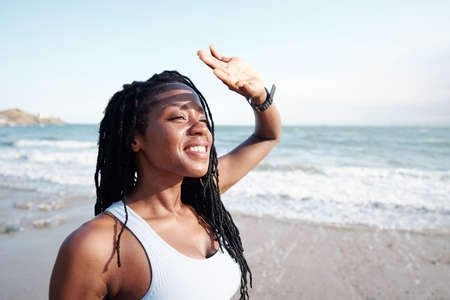 Cheerful young woman covering her eyes with hands from bright sunlight when looking at seascapeの写真素材