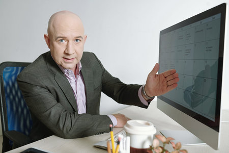 Portrait of serious mature CEO in gray suit sitting at table and telling about his timetable while pointing at computer screen in officeの写真素材
