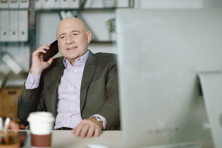Smiling confident bald Caucasian executive in gray suit sitting at table in office and calling by...