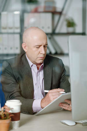 Concentrated mature analyst with bald head sitting at desk and working with data in officeの写真素材