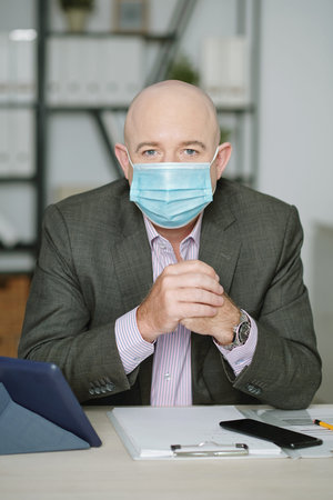 Portrait of mature bald business executive in mask sitting at desk with clipboard and gadgets and...