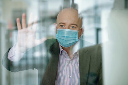 Behind glass view of sad mature businessman in mask touching glass box while being in isolation...