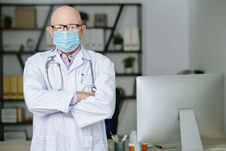Portrait of confident experienced mature Caucasian doctor in eyeglasses and mask standing with crossed arms in modern officeの写真素材