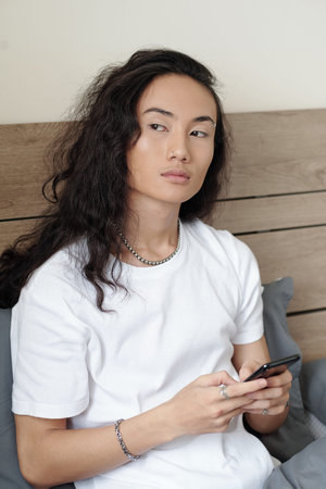 Serious long-haired Vietnamese guy with wavy black hair sitting on bed and looking away while...