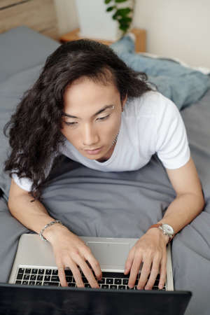 Serious young Vietnamese guy with wavy hair wearing earring in eye brow lying on bed and typing on laptopの写真素材