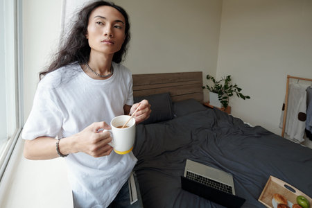 Pensive young Vietnamese man in wavy hair standing at window and stirring sugar in coffee cup, he...