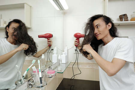 Smiling young Vietnamese man with long hair standing in front of mirror and using hairdryer in bathroomの写真素材