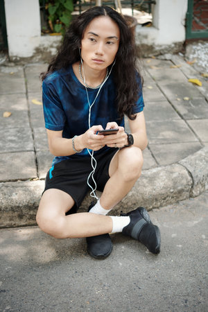 Serious handsome young Asian man with curly hair sitting on curb and listening to audio message in earphones outdoorsの写真素材