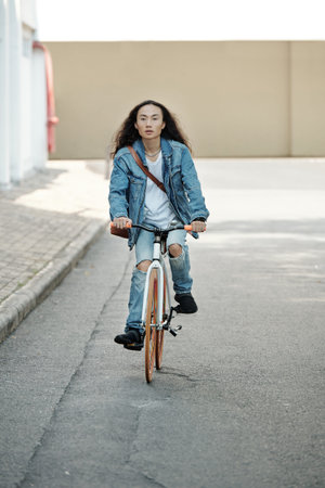 Active young Vietnamese man with long hair wearing jeans with holes on knees looking around while...