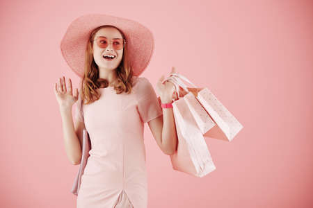 Portrait of excited young woman in hat and sunglasses holding shopping bags and waving with handの写真素材