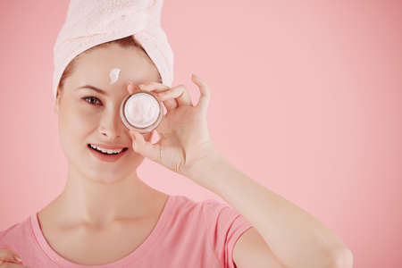 Smiling pretty young woman showing lifting and brightening face cream she is applying after morning showerの写真素材
