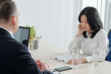 Serious young businesswoman looking at her cv when talking to HR manager at office tableの写真素材