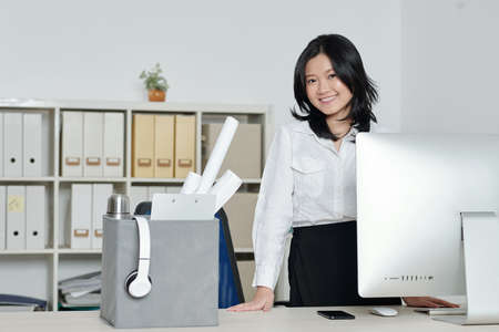 Happy beautiful young woman standing at her new office desk with computer and box of belongingsの写真素材