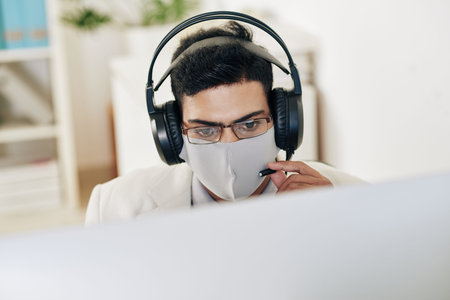 Serious technical support operator in medical mask and headphones answering phone call of customer