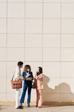 Group of business people standing oudoors in protective masks and discussing various documents and...