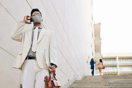 Handsome young businessman in white suit and medical mask walking down the stairs leaving office building and talking on phone with colleague or business partnerの写真素材