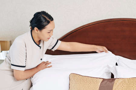 Young maid changing bed sheets and preparing hotel room for next guestの写真素材