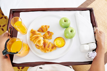Maid carrying tray with breakfast to hotel room, it consist of fresh croissants, apples, and glasses of juice, view from aboveの写真素材