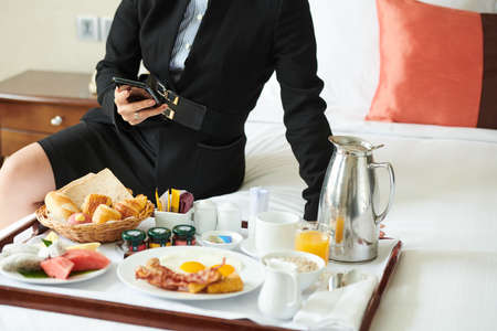 Young businesswoman sitting on hotel bed, eating breakfast and checking social media on her smartphoneの写真素材