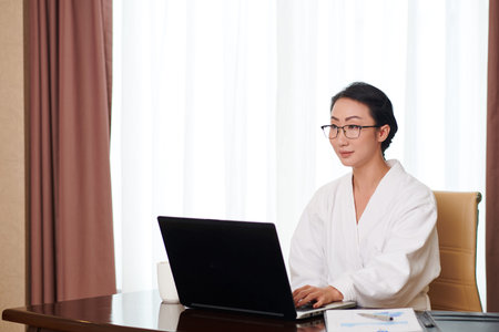 Portrait of young pretty businesswoman in bathrobe and glasses working on laptop at homeの写真素材