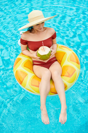 Young woman in straw hat sitting on inflatable ring in swimming pool and drinking coconut cocktailの写真素材