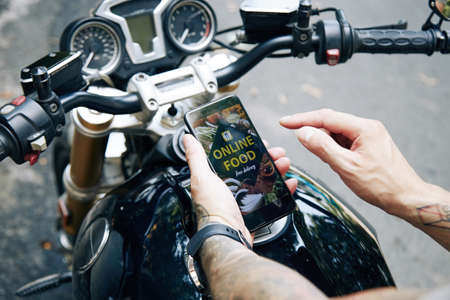Hands of man sitting on motorcycle and using mobile application to order food delivery from local cafeの写真素材