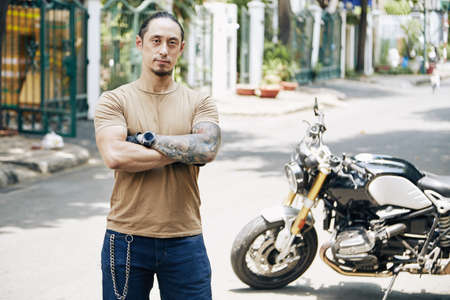 Portrait of confident serious fit man standing outdoors in front of his motorcycleの写真素材