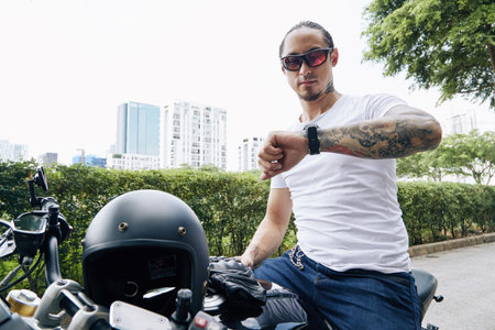 Man in sunglasses and white t-shirt sitting on motorcycle or scooter and checking time or...
