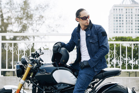 Stylish man in sunglasses, jeans and denim jacket sitting on motorcycle, taking off helmet and...