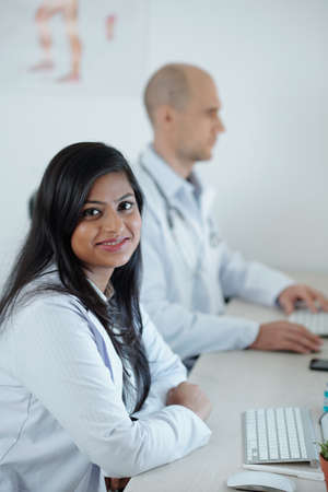 Smiling young doctor sitting at desk next to his colleague and working on laptop, filling patients dataの写真素材