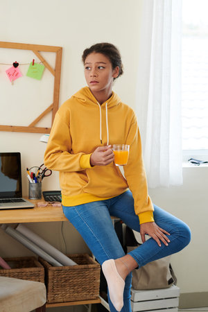 Sad mixed race teenage girl in casual outfit sitting on desk with supplies and laptop and drinking...