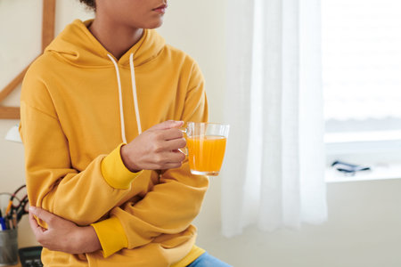 Close-up of mixed race girl in yellow hoodie sitting in room and holding mug of juice