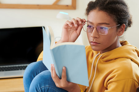 Serious mixed race school girl sitting at desk with laptop and adjusting eyeglasses while reading...