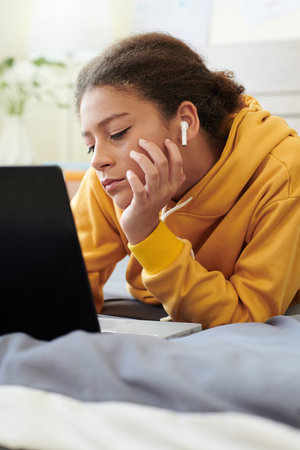 Boring mixed race teenage girl in ear buds and yellow hoodie lying on bed and using laptop while...