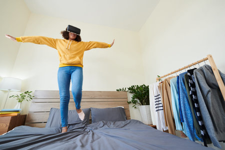 Below view of cheerful carefree mixed race girl in virtual reality goggles jumping with outstretched...