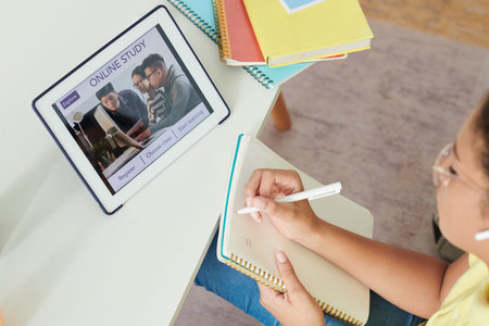 Above view of concentrated girl sitting at table with colorful workbooks and making notes on online...