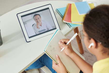 Over shoulder view of mixed race girl in ear buds making notes in workbook while watching webinar on...