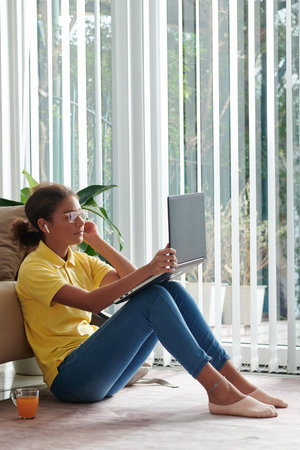 Content mixed race girl in wireless ear buds and eyeglasses sitting on floor with mug of juice and...