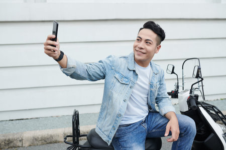 Positive young Vietnamese man in denim clothes sitting on moped and taking selfie on smartphone...