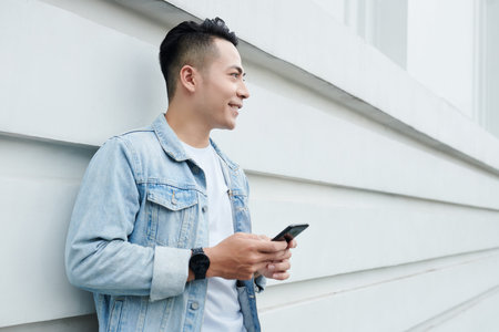 Smiling handsome young Vietnamese man in denim jacket standing with smartphone against building wall