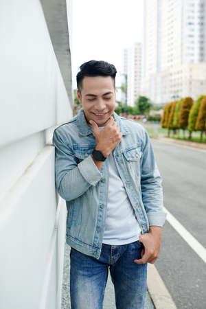 Smiling handsome young Vietnamese man in wristwatch leaning on building wall and touching chin on streetの写真素材