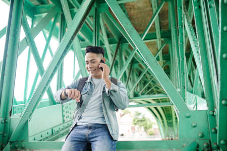 Smiling young Asian man with satchel sitting on metal girder and checking time while calling by phone under bridgeの写真素材