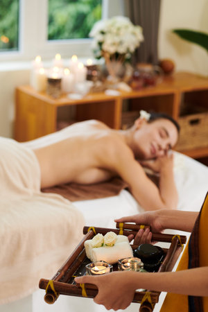 Close-up of therapist holding tray with candles and aroma towels while woman relaxing on sofa in the...